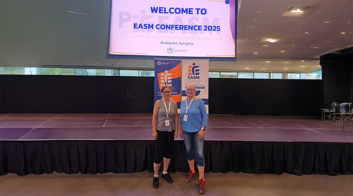 Zwei Teilnehmerinnen der FH Kufstein Tirol stehen lächelnd vor einer Bühne mit EASM-Banner und Konferenzschild bei der EASM Conference 2025 in Budapest, Ungarn.