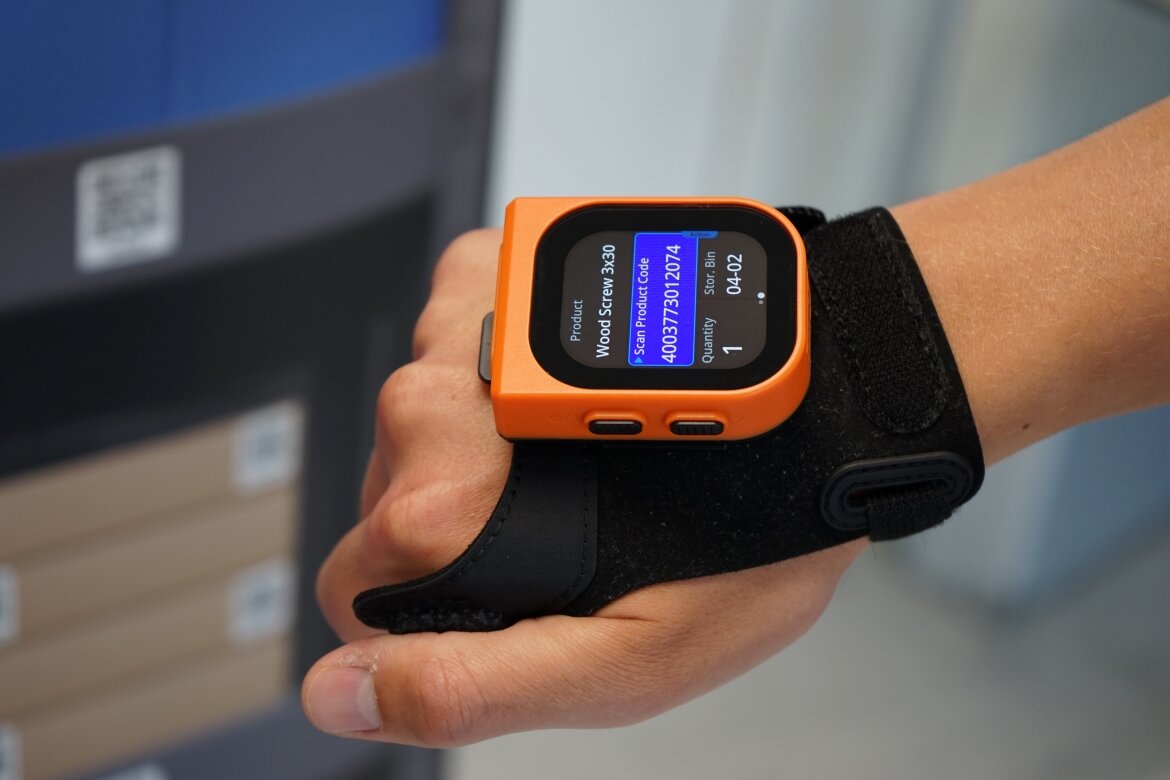 Nahaufnahme einer Hand mit einem tragbaren ProGlove-Scanner am Handrücken. Auf dem kleinen Display ist der Lagerplatz sowie ein Produktcode für einen Holzschraubenartikel angezeigt.