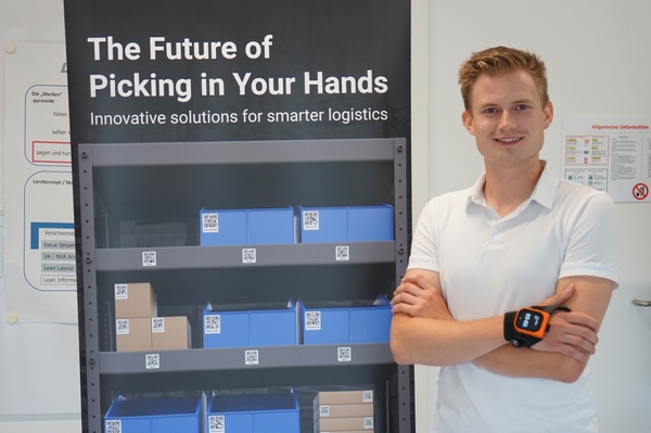 Ein junger Mann in weißem Poloshirt steht mit verschränkten Armen vor einem Roll-up mit dem Titel „The Future of Picking in Your Hands“. Am Handgelenk trägt er einen ProGlove-Scanner. | © FH Kufstein Tirol