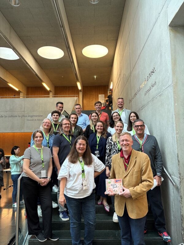 Gruppenfoto von rund 20 Personen auf einer Treppe in einem modernen Innenraum mit Betonwänden und Lichtringen an der Decke. Im Vordergrund stehen zwei Frauen und ein Mann mit einer Schachtel Mozartkugeln, alle lachen in die Kamera. Viele tragen Namensschilder und grüne INNoCamp-Lanyards.