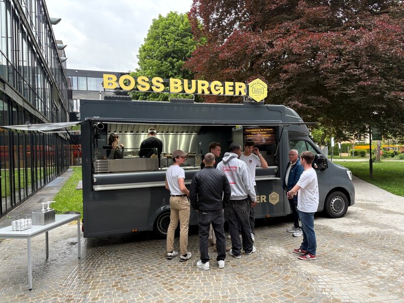 Mehrere Personen stehen vor einem grauen Foodtruck mit gelbem „BOSS BURGER“-Schriftzug. Im Truck bereiten zwei Mitarbeitende Speisen zu. Die Szene spielt sich auf dem gepflasterten Außenbereich eines modernen Campusgebäudes ab, umgeben von Bäumen und Glasfassaden.