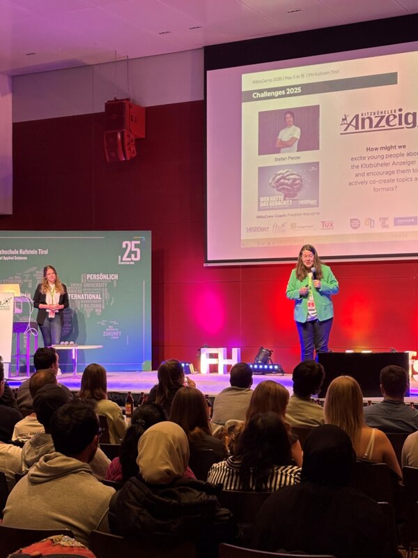 Eine Frau in grünem Blazer steht auf einer Bühne und spricht vor einem Publikum. Im Hintergrund ist eine große Leinwand mit einer Präsentationsfolie zur „Challenge 2025“ des Kitzbüheler Anzeigers zu sehen. Das Publikum sitzt im dunkleren Vordergrund.