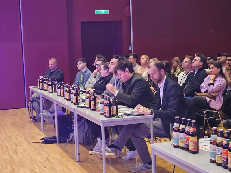 Mehrere Jury-Mitglieder sitzen konzentriert an Tischen bei der INNoCamp 2025. Vor ihnen stehen viele Flaschen einer Limonadenmarke. Im Hintergrund verfolgt das Publikum gespannt die Präsentation.