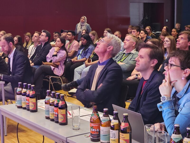Jury-Mitglieder und Publikum sitzen aufmerksam in einem Vortragssaal beim INNoCamp 2025. Die Juroren verfolgen gespannt eine Präsentation, auf den Tischen stehen zahlreiche Getränkeflaschen.