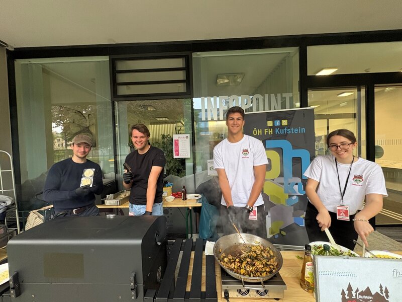 Vier junge Menschen stehen an einem Kochstand vor dem Infopoint der FH Kufstein und bereiten gemeinsam Gemüsegerichte zu. Im Hintergrund ist ein ÖH-Banner sichtbar.