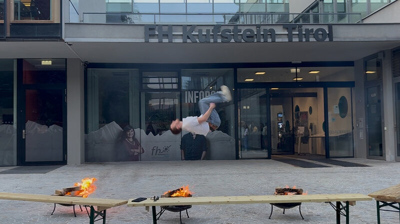 Ein junger Mann macht einen Rückwärtssalto zwischen zwei Feuerschalen vor dem Haupteingang der FH Kufstein Tirol. Im Hintergrund ist das Gebäude mit Glasfassade und das FH-Logo zu sehen.