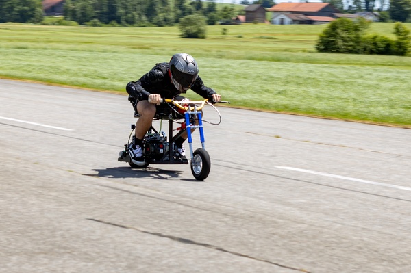 Ein junger Mann mit Schutzkleidung und Helm fährt in geduckter Haltung ein selbstgebautes Mini-Bike auf einer Landstraße. Das Fahrzeug ist extrem klein und liegt tief über dem Asphalt. | © FH Kufstein Tirol