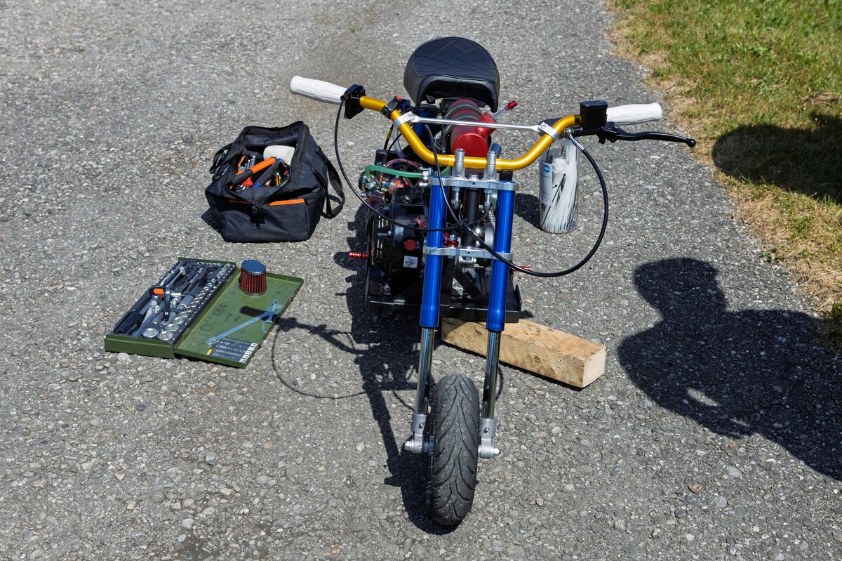 Detailansicht des Tiny Torquer Mini-Bikes mit Werkzeug Frontale Nahaufnahme eines kleinen Mini-Bikes mit sichtbarem Motor, goldfarbenem Lenker und blauen Gabelrohren. Daneben liegen Werkzeugkoffer, ein Luftfilter und eine Tasche mit Schraubwerkzeug auf dem Asphalt.