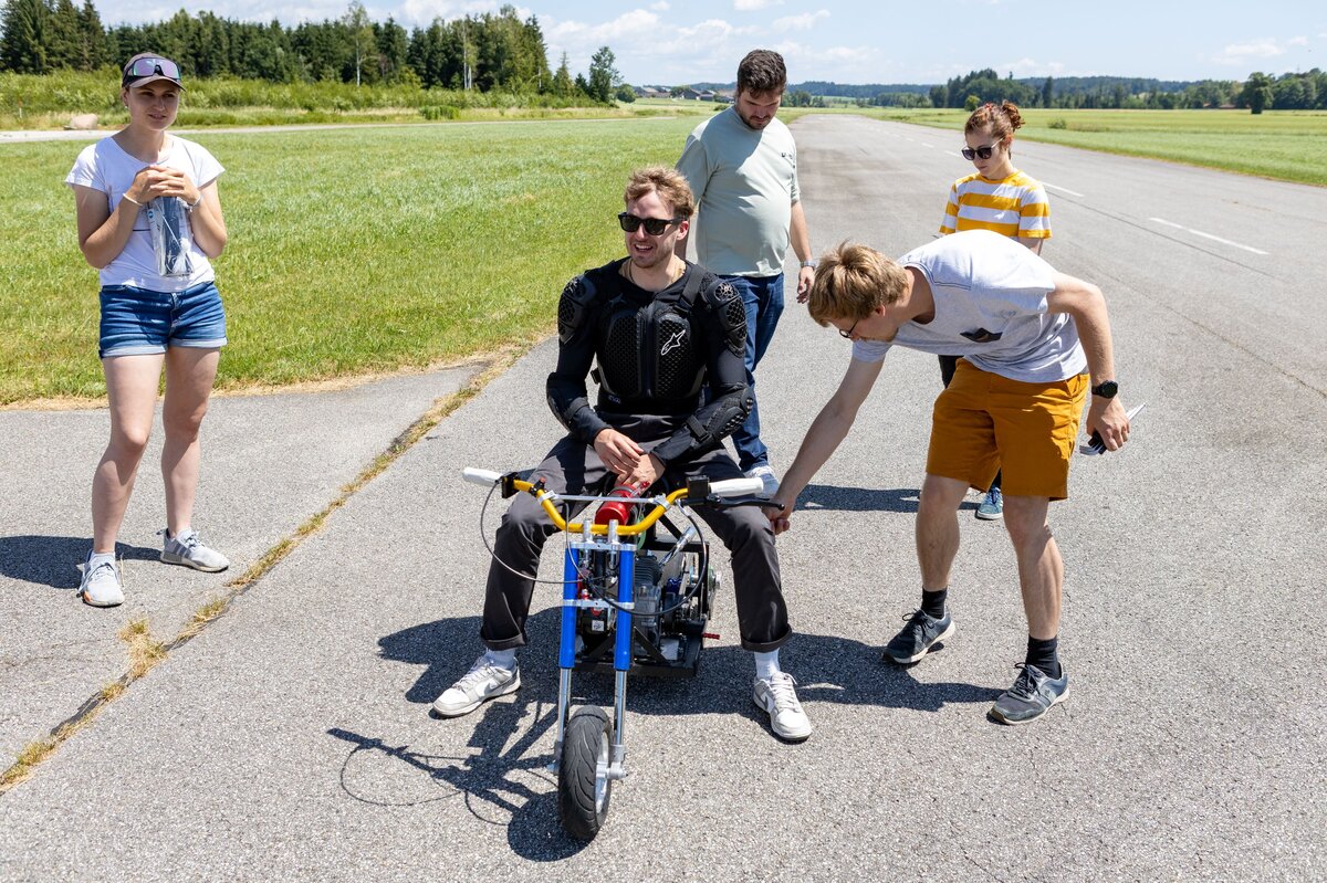 Eine Gruppe junger Erwachsener überprüft ein kleines motorisiertes Mini-Bike auf einer Landebahn in Vogtareuth. Der Fahrer trägt Schutzkleidung und sitzt noch auf dem Bike, während ein Teammitglied sich zum Bike herunterbeugt.