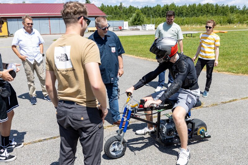 Ein junger Mann in Schutzkleidung und Helm sitzt auf einem selbstgebauten Mini-Bike, umgeben von mehreren Personen, die sich austauschen und die Technik begutachten. Die Szene spielt sich auf einem sonnigen Testgelände in Vogtareuth ab.