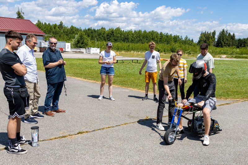 Eine Gruppe Studierender und Betreuer:innen steht auf einem Flugplatz in Vogtareuth um ein selbstgebautes Mini-Bike. Eine Person sitzt noch mit Helm und Schutzkleidung auf dem Fahrzeug, während ein Teammitglied am Lenker eine Justierung vornimmt. Die restlichen Personen beobachten die Szene aufmerksam.