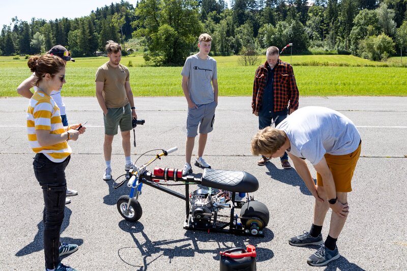 Sechs Personen stehen auf einem Flugplatz um ein technisches Mini-Bike. Eine Person beugt sich über das Fahrzeug und inspiziert es, während die anderen beobachten oder in Gespräche vertieft sind. Im Hintergrund ist eine grüne Wiese mit Windsack und Bäumen zu sehen.