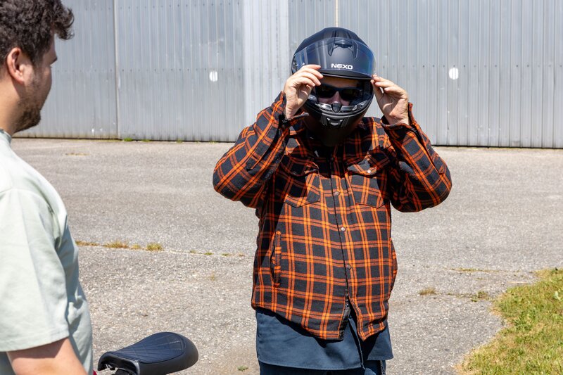 Ein Mann in orange-schwarz karierter Jacke setzt sich auf einem Flugplatz einen schwarzen Motorradhelm auf. Im Vordergrund ist ein Teil des Mini-Bikes zu sehen, daneben steht eine weitere Person.