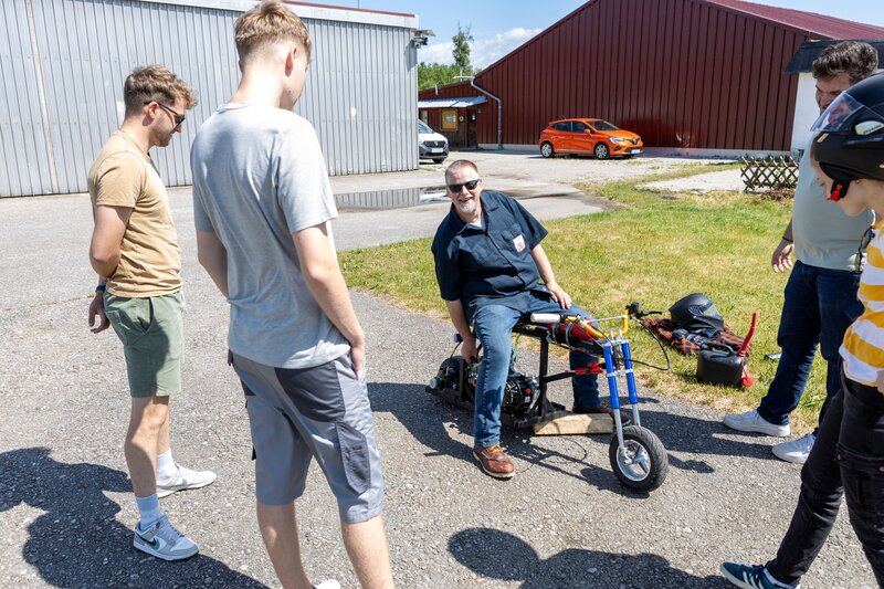 Prof. Schmid sitzt auf einem Mini-Bike und spricht mit einer Gruppe Studierender auf einem Flugplatz. Neben dem Bike liegen Helm und Werkzeug. Im Hintergrund sind Hallen und ein orangefarbenes Auto zu sehen.
