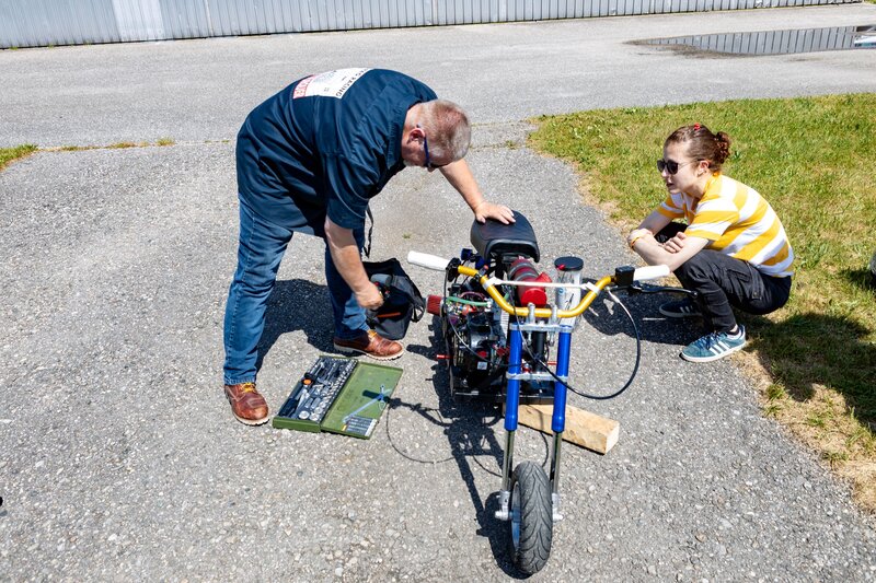 Prof. Schmid beugt sich über ein Mini-Bike und greift in seine Werkzeugtasche, während Studentin Sofia Tanoni daneben in der Hocke sitzt und zuschaut. Werkzeug liegt auf dem Boden, im Hintergrund sind Asphalt und Gras zu sehen.