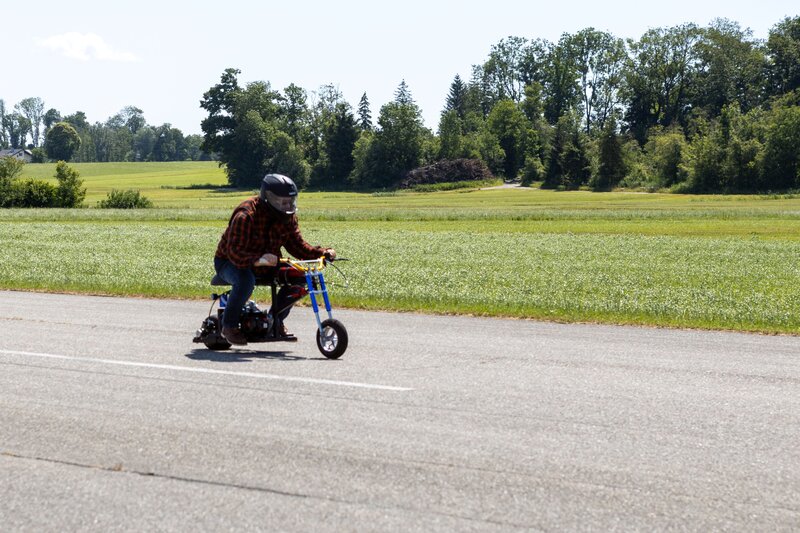 Prof. Schmid fährt mit Schutzkleidung und Helm ein kleines Mini-Bike über eine Landebahn, umgeben von grünen Wiesen und Bäumen im Hintergrund.