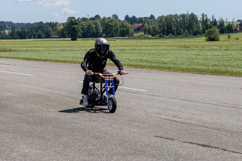 Alexander Wohlfart fährt auf einem Mini-Bike mit Schutzkleidung und Helm auf einer asphaltierten Teststrecke, umgeben von grünen Wiesen und Bäumen im Hintergrund.