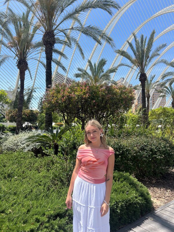 Maya in einem grünen Garten mit Palmen und Pflanzen; im Hintergrund moderne Architektur mit weißen, geschwungenen Balken vor blauem Himmel.