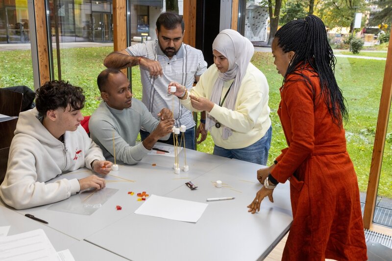 Eine diverse Gruppe von Studierenden arbeitet gemeinsam an einem Turm aus Spaghetti, Marshmallows und Gummibärchen.