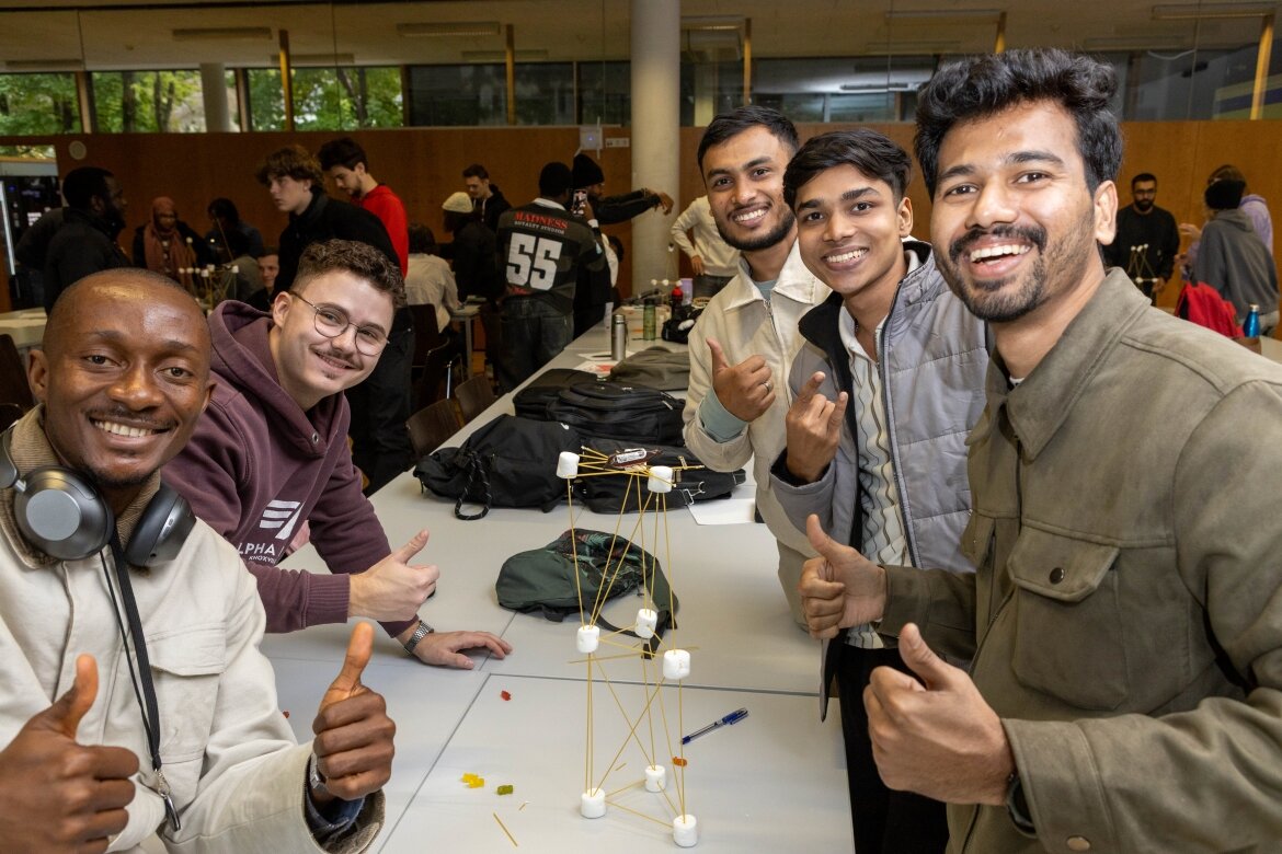 Erfolgreiches Team präsentiert fertigen Marshmallow-Turm Eine Gruppe lachender Studierender zeigt stolz ihren fertigen Turm aus Marshmallows und Spaghetti im Workshop.