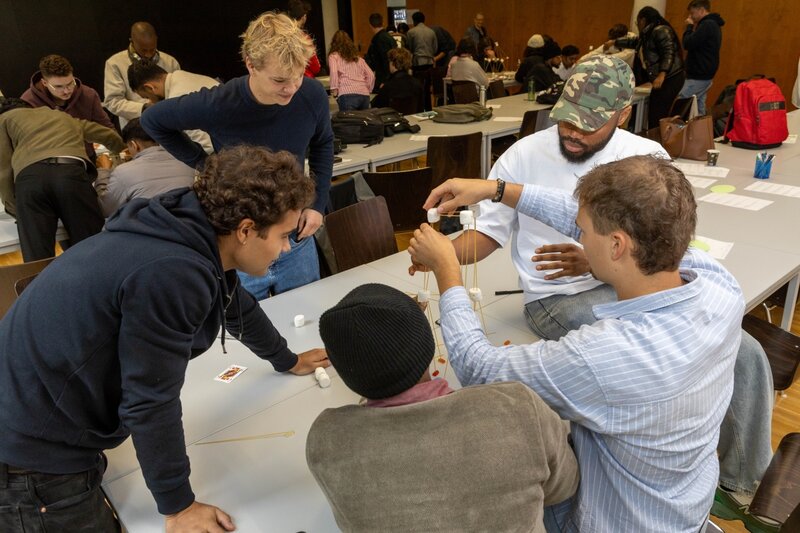 Studierende arbeiten konzentriert im Team an einem Turm aus Marshmallows und Spaghetti während eines Workshops.