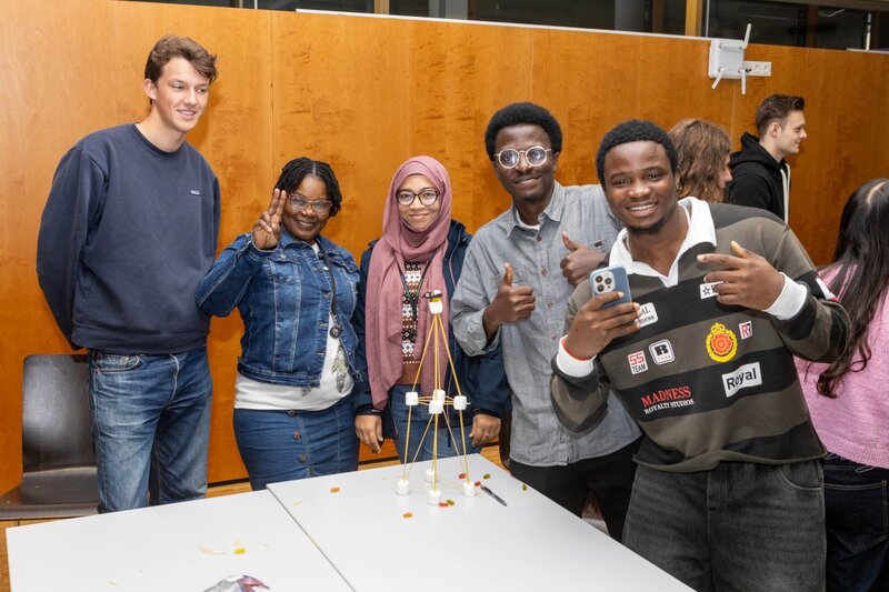 Sechs Studierende posieren lachend hinter ihrem selbst gebauten Marshmallow-Turm und zeigen Daumen hoch oder Friedenszeichen.