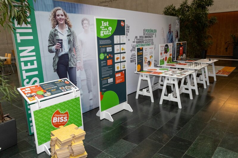 Verschiedene interaktive Stationen der Ausstellung „Gewissensbiss“ in der Aula der FH Kufstein Tirol. Die Tafeln und Tische informieren spielerisch über Lebensmittelverschwendung, Haltbarkeit und Geschmack.