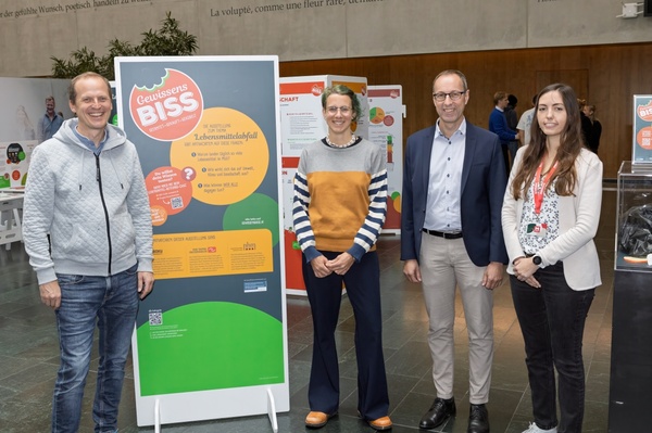 Vier Personen stehen neben einem großen Informationsplakat zur Ausstellung „Gewissensbiss“ in der Aula der FH Kufstein Tirol. Die Ausstellung thematisiert Lebensmittelabfall und nachhaltigen Konsum. Im Hintergrund sind weitere interaktive Stationen sichtbar. | © FH Kufstein Tirol