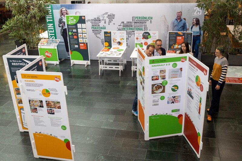 Blick von oben auf die Ausstellung „Gewissensbiss“ in der Aula der FH Kufstein Tirol. Die Ausstellung umfasst Informationstafeln und Mitmachstationen zu Themen wie Lebensmittelabfall, Konsum und Haltbarkeit. Einige Besucher:innen betrachten interaktiv die Inhalte.