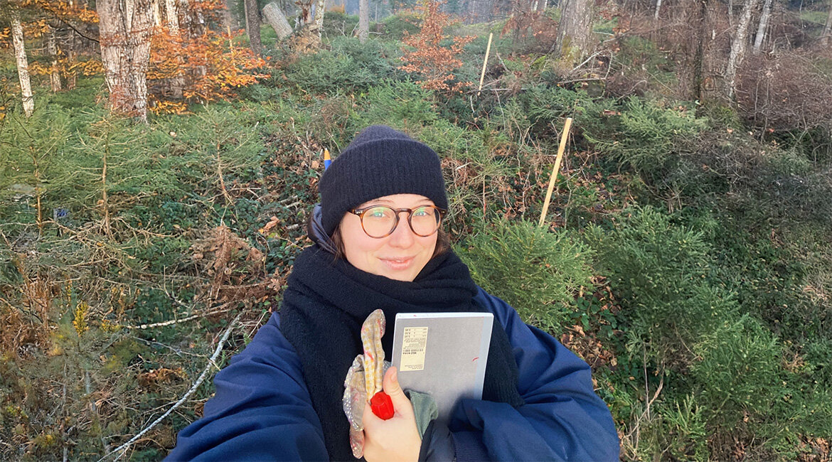 Nina Oberreiter trägt warme Kleidung und steht mit Notizbuch und Handschuhen im Stadtwald Kufstein. Im Hintergrund sind Markierungsstäbe und junge Bäume zu sehen.