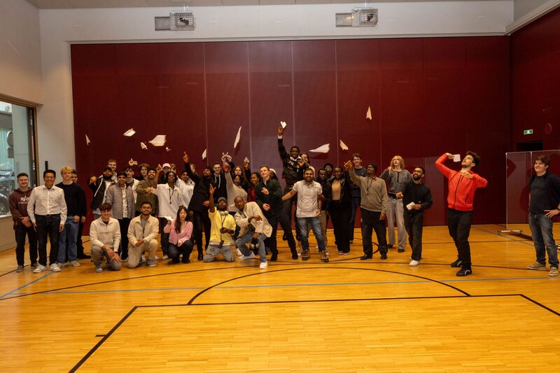Personen stehen in der Halle zusammen und werfen die Papierflieger hoch.