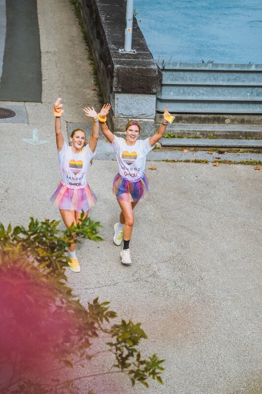 Zwei Personen laufen in einer Gasse nebeneinander und winken in die Kamera.