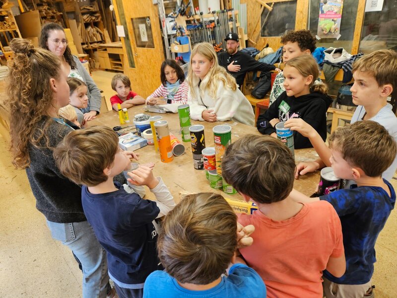 Kinder stehen um einen Werktisch und schauen zur Workshopleiterin.