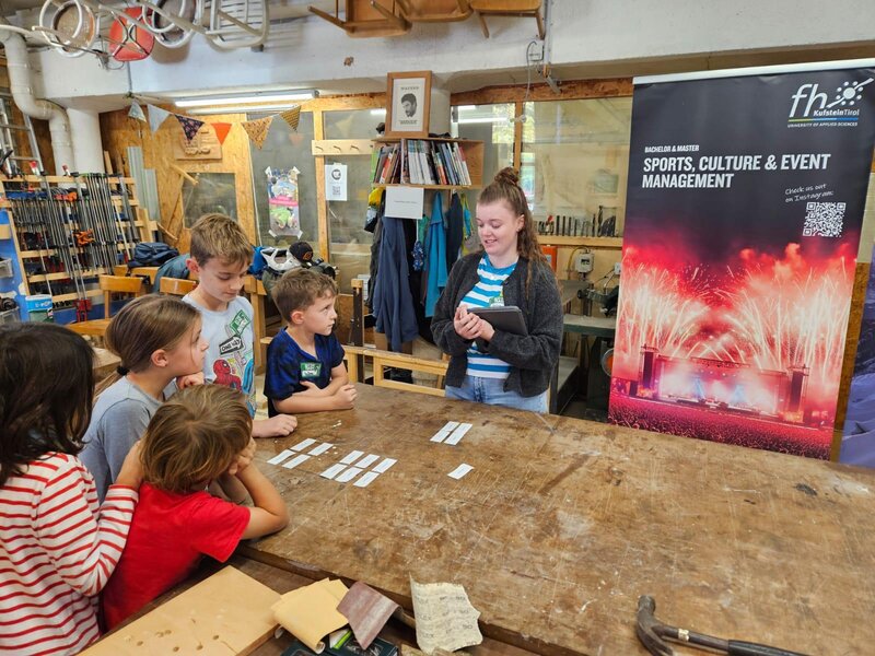 Kinder stehen um einen Werktisch und schauen zur Kursleiterin.