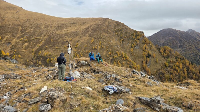 Eine Person steht bei der Wetterstation und zwei weitere Personen arbeiten wieiter entfernt.