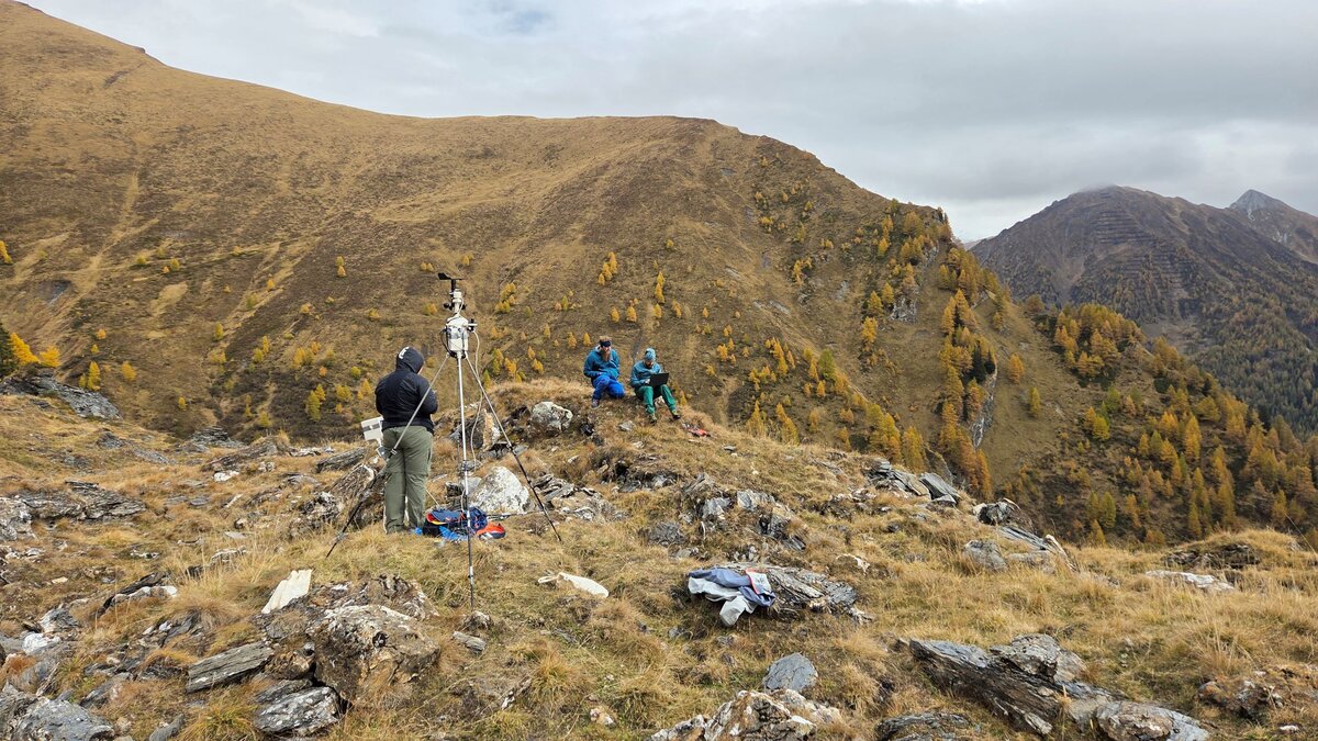 Eine Person steht bei der Wetterstation und zwei weitere Personen arbeiten wieiter entfernt.
