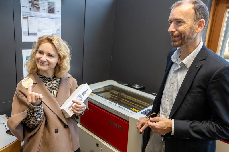 Zwei Personen stehen nebeneinander und die linke Person hält einen runden Holzchip nach oben.