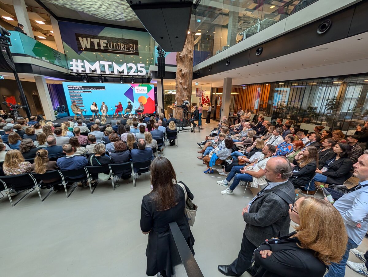 Personen sitzen in einer Aula zur Bühne gerichtet.