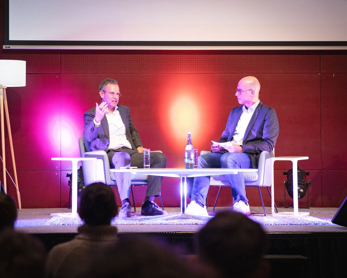 Zwei Personen sitzen auf einer Bühne auf Stühlen vor einem Tisch.