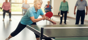 Eine ältere Dame steht mit gebeugten Knien mit einem Tischtennis-Schläger vor einer Spielplatte.