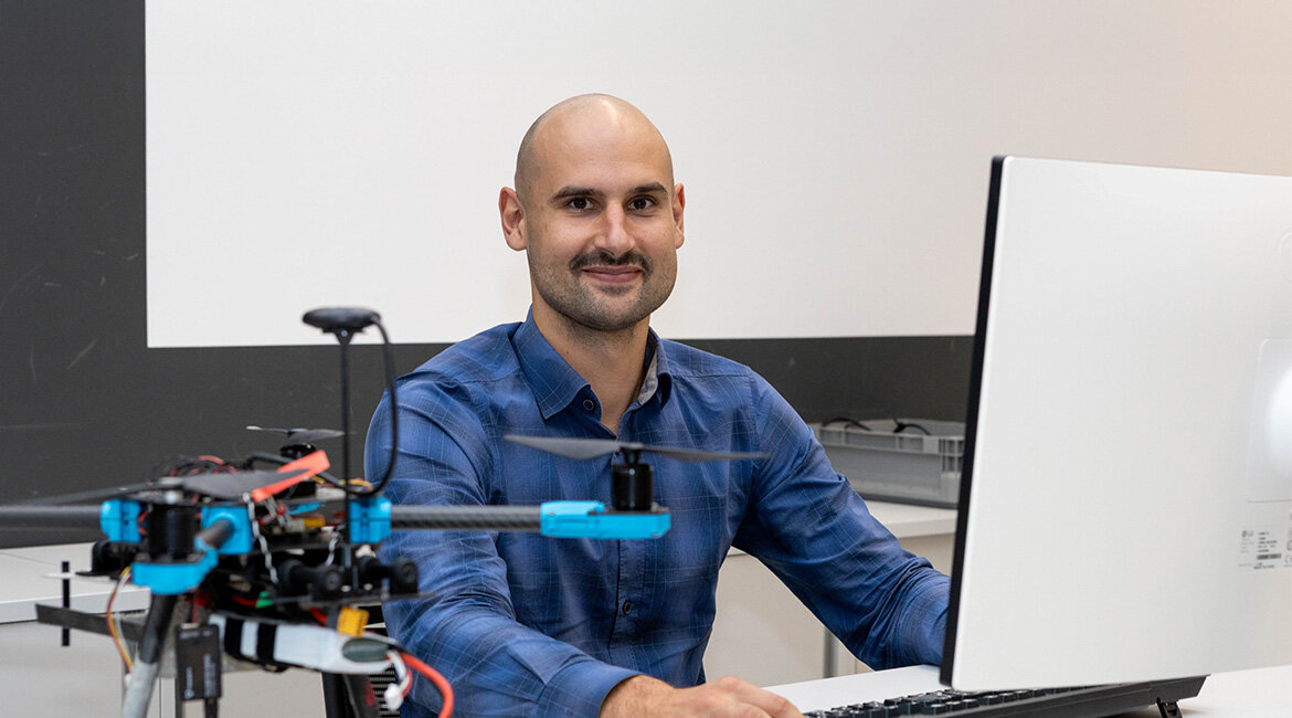 Julian Bialas sitzt an einem Computer, im Vordergrund ist eine Drohne auf seinem Schreibtisch zu sehen.