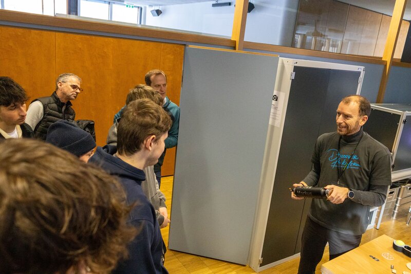 Personen stehen in einem Raum und sind dem vortragenden Dozent zugewendet.