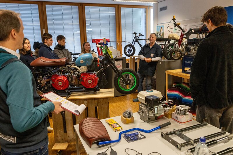 Personen stehen in einer Werkstatt mit kleinen Motorrädern auf Regalen.