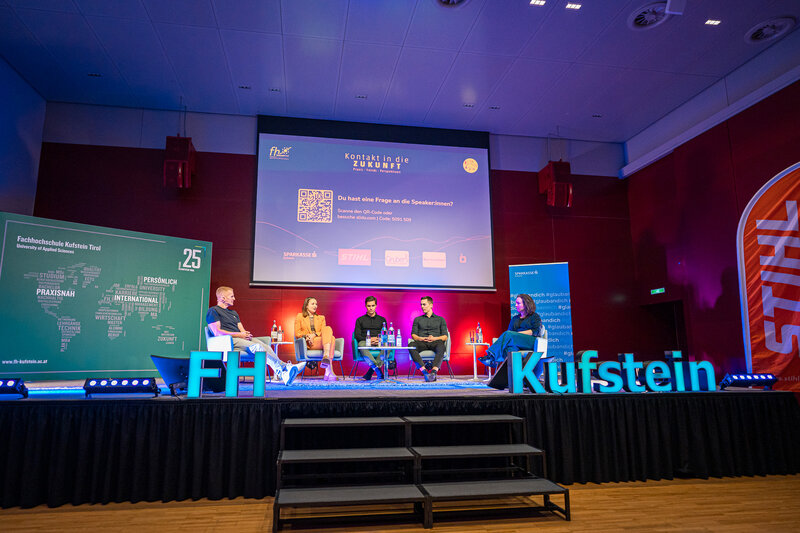 Personen sitzen auf einer Buehne mit verschiedenen Bildschirmen.