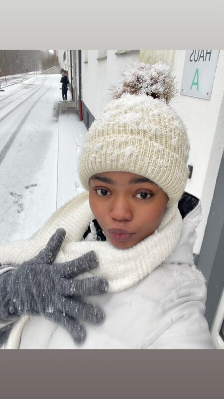 Person steht im Schnee und blickt in die Kamera und hat auf ihrer beigen Mütze Schnee.