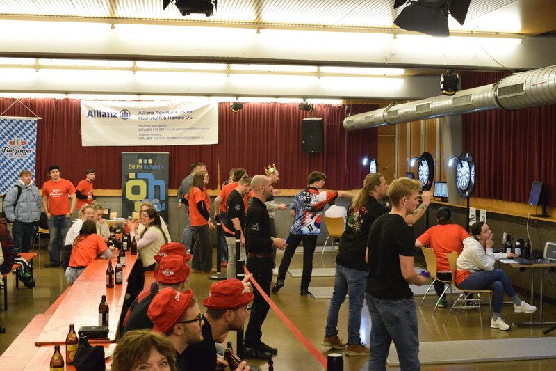 Personen stehen in einer Halle und am Rand sind Dartscheiben aufgestellt.