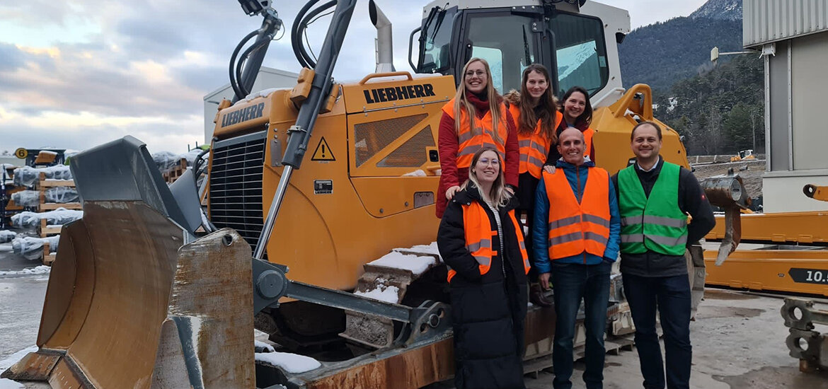 Praxisprojekt-Team-Liebherr-Werk-Telfs | © FH Kufstein Tirol Das Praxisprojekt-Team mit Projektauftraggeber Tobias Apperl (1.v.r.) vor einem Liebherr-Bagger am schneebedeckten Baugelände. | © FH Kufstein Tirol