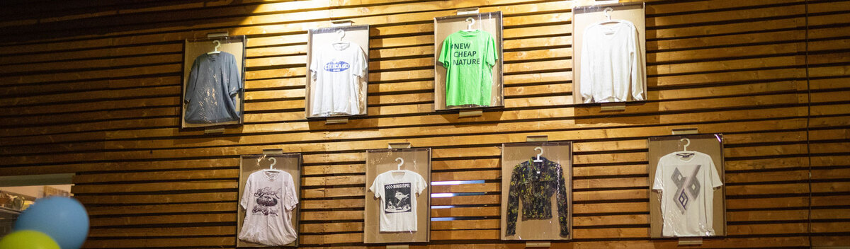 T-Shirts-Holzwand-Modegeschäft | © FH Kufstein Tirol Auswahl an T-Shirts, präsentiert an einer Holzwand in einem Modegeschäft, beleuchtet durch warmes Licht von oben. | © FH Kufstein Tirol