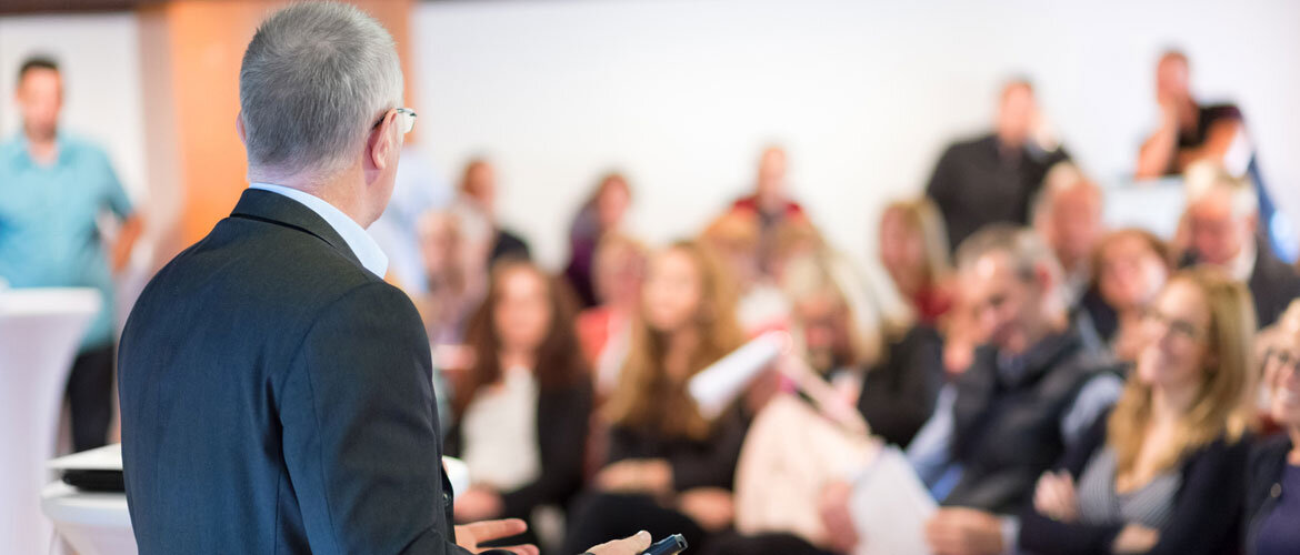 Redner-im-Austausch-mit-Publikum-bei-Konferenzveranstaltung Ein Redner, der mit dem Rücken zur Kamera steht, spricht vor einem Publikum in einem Konferenzraum.
