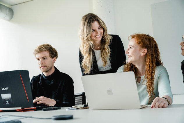 Arbeiten_am_Laptop | © FH Kufstein Tirol Drei Personen sitzen an einem Tisch und arbeiten an ihren Laptops, während eine stehende Frau etwas mit einer sitzenden Frau bespricht. | © FH Kufstein Tirol